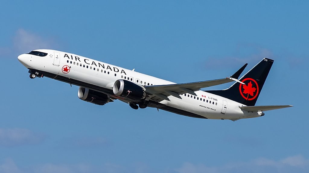 Air Canada Boeing 737 MAX‑8 aircraft in mid‑flight with clear sky in background, featuring white fuselage, black tail, and red maple leaf logo.