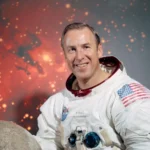 Official NASA portrait of astronaut Jim Lovell in a white Apollo-era spacesuit, smiling with an American flag patch, posed against a lunar-themed backdrop.