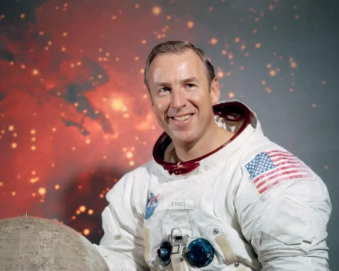 Official NASA portrait of astronaut Jim Lovell in a white Apollo-era spacesuit, smiling with an American flag patch, posed against a lunar-themed backdrop.