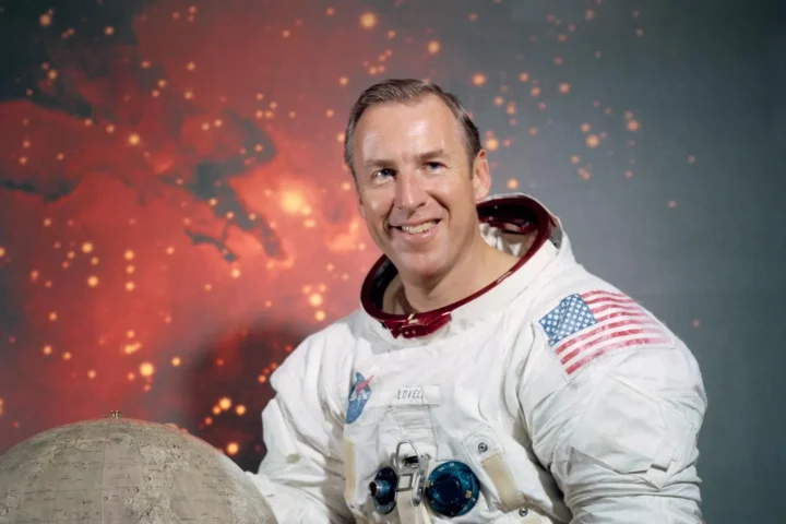 Official NASA portrait of astronaut Jim Lovell in a white Apollo-era spacesuit, smiling with an American flag patch, posed against a lunar-themed backdrop.