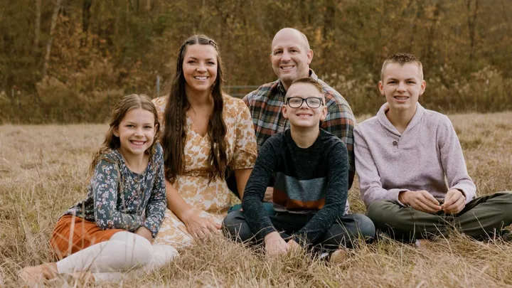 Family portrait of Ariean Fabrizio Colton with her husband and three children sitting together in a grassy field, smiling at the camera.
