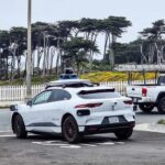 White Waymo Jaguar I‑Pace autonomous car with roof sensors parked on Point Lobos Avenue in San Francisco, with a white fence and trees in the background