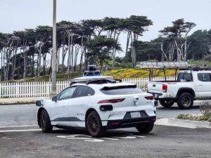 White Waymo Jaguar I‑Pace autonomous car with roof sensors parked on Point Lobos Avenue in San Francisco, with a white fence and trees in the background