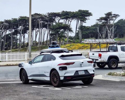 White Waymo Jaguar I‑Pace autonomous car with roof sensors parked on Point Lobos Avenue in San Francisco, with a white fence and trees in the background