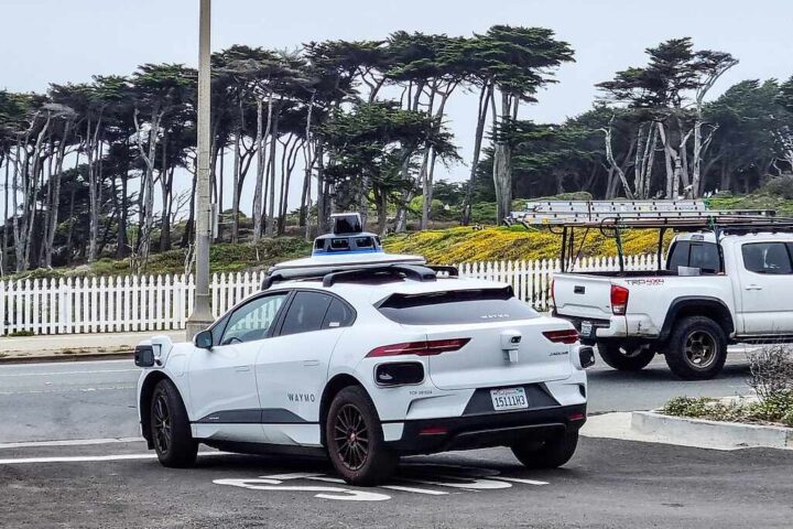 White Waymo Jaguar I‑Pace autonomous car with roof sensors parked on Point Lobos Avenue in San Francisco, with a white fence and trees in the background