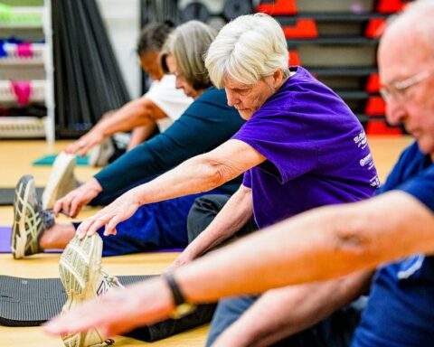 Group of older adults seated on mats in a gym, stretching toward their feet with exercise shelves in the background