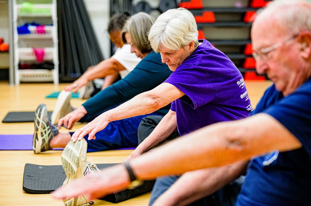 Group of older adults seated on mats in a gym, stretching toward their feet with exercise shelves in the background