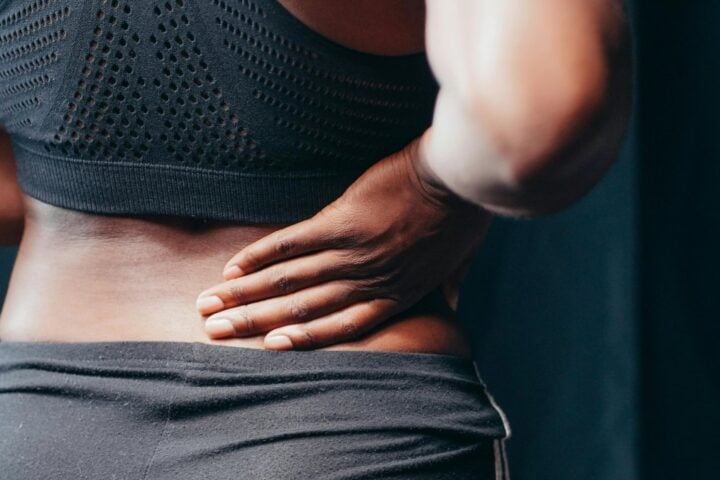 Person in athletic wear holding their lower back in pain, shown from behind with focus on the torso and hand placement indicating back discomfort.