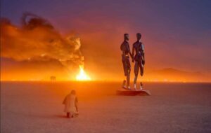 Two chrome figures from Michael Benisty’s “Broken Together” sculpture stand before the burning temple at dusk on the Black Rock Desert playa during Burning Man 2019.