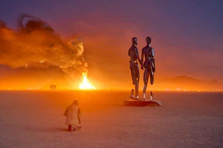 Two chrome figures from Michael Benisty’s “Broken Together” sculpture stand before the burning temple at dusk on the Black Rock Desert playa during Burning Man 2019.