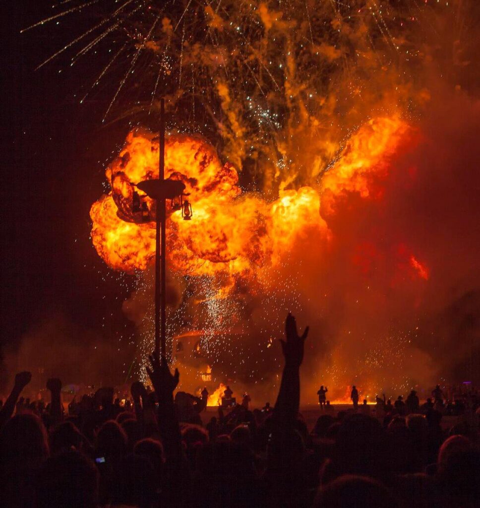 Silhouetted crowd with raised hands watches a massive wooden effigy engulfed in bright orange flames at night during Burning Man 2012 in the Black Rock Desert.