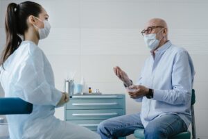 Elderly man speaks with a healthcare professional in a white medical gown and gloves in a clinic; medical cabinet with bottles, containers, and cotton swabs in the background.