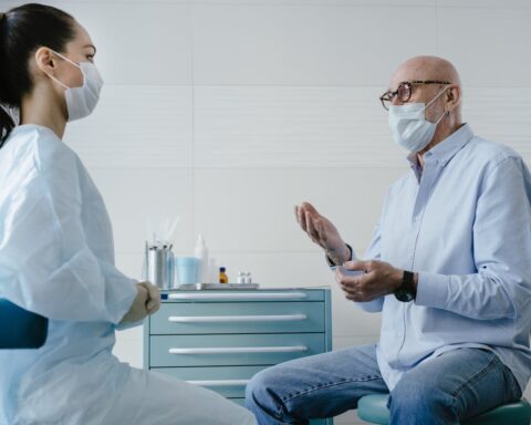 Elderly man speaks with a healthcare professional in a white medical gown and gloves in a clinic; medical cabinet with bottles, containers, and cotton swabs in the background.