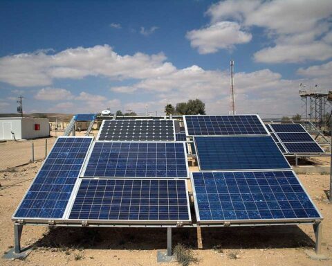 Multiple rows of blue photovoltaic solar panels mounted on metal frames in an outdoor testing facility under clear blue skies with some buildings visible in the background