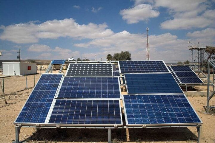 Multiple rows of blue photovoltaic solar panels mounted on metal frames in an outdoor testing facility under clear blue skies with some buildings visible in the background