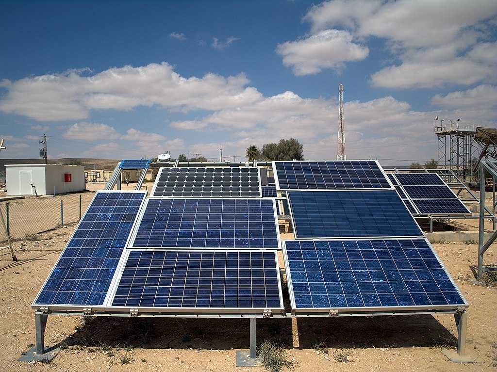 Multiple rows of blue photovoltaic solar panels mounted on metal frames in an outdoor testing facility under clear blue skies with some buildings visible in the background