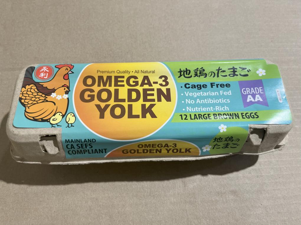 Carton of 12 large brown Omega-3 Golden Yolk eggs with colorful packaging featuring a chicken illustration, labeled as cage-free, vegetarian-fed, antibiotic-free, and Grade AA.