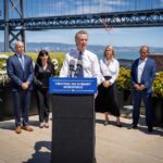 Governor Gavin Newsom at an outdoor podium flanked by representatives from Adobe, Google, IBM, and Microsoft, with the San Francisco–Oakland Bay Bridge visible in the background.