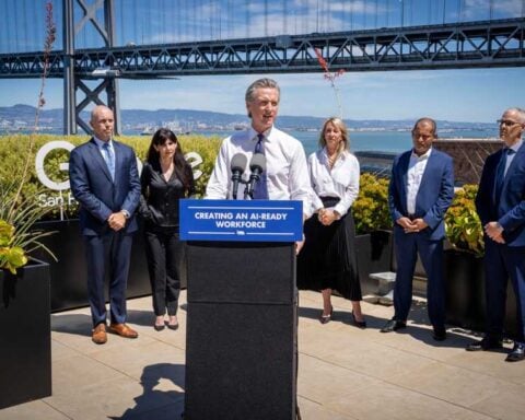 Governor Gavin Newsom at an outdoor podium flanked by representatives from Adobe, Google, IBM, and Microsoft, with the San Francisco–Oakland Bay Bridge visible in the background.