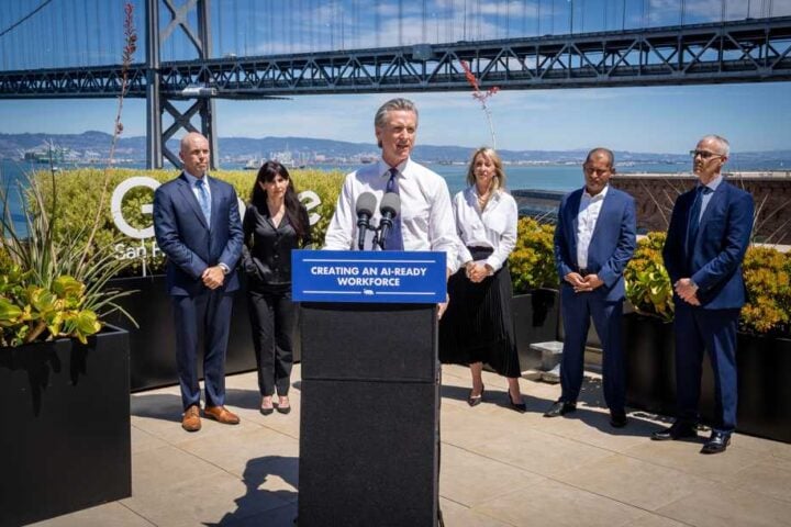 Governor Gavin Newsom at an outdoor podium flanked by representatives from Adobe, Google, IBM, and Microsoft, with the San Francisco–Oakland Bay Bridge visible in the background.