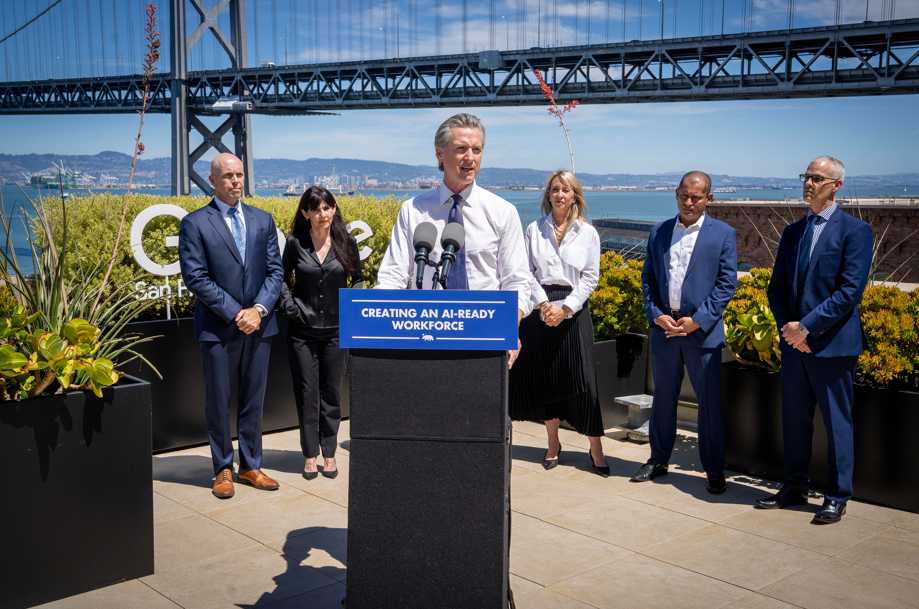 Governor Gavin Newsom at an outdoor podium flanked by representatives from Adobe, Google, IBM, and Microsoft, with the San Francisco–Oakland Bay Bridge visible in the background.