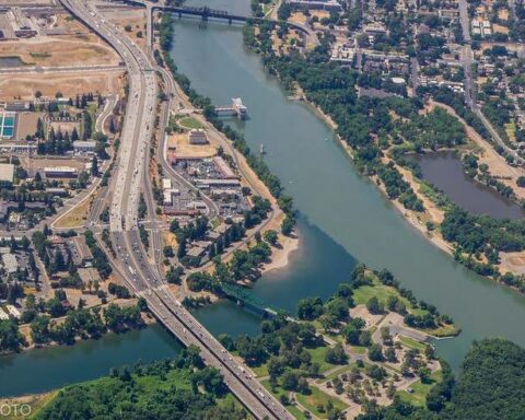 Aerial view of the American River meeting the Sacramento River at Discovery Park in Sacramento, with surrounding roads, buildings, and green spaces visible.