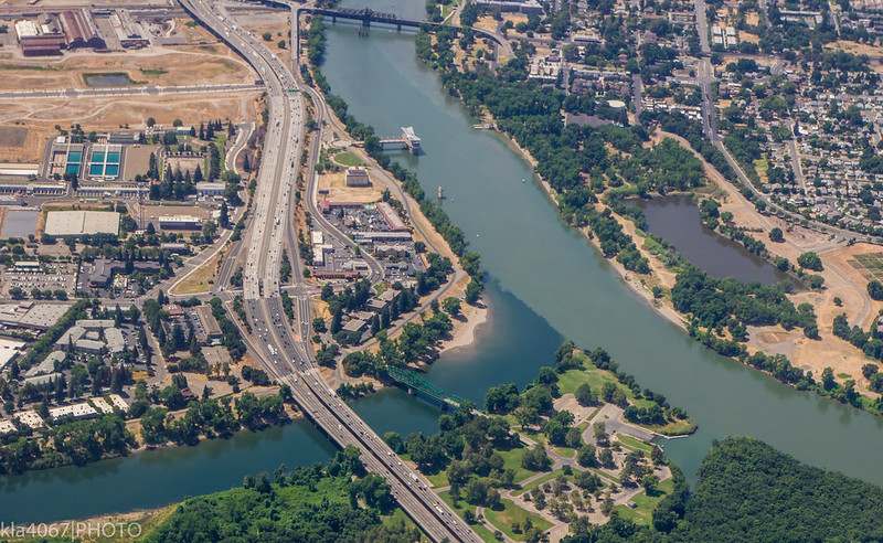 Aerial view of the American River meeting the Sacramento River at Discovery Park in Sacramento, with surrounding roads, buildings, and green spaces visible.
