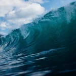 Close-up perspective of a large turquoise ocean wave curling with white foam against a blue sky with clouds, demonstrating the immense power and scale of tsunami waves.