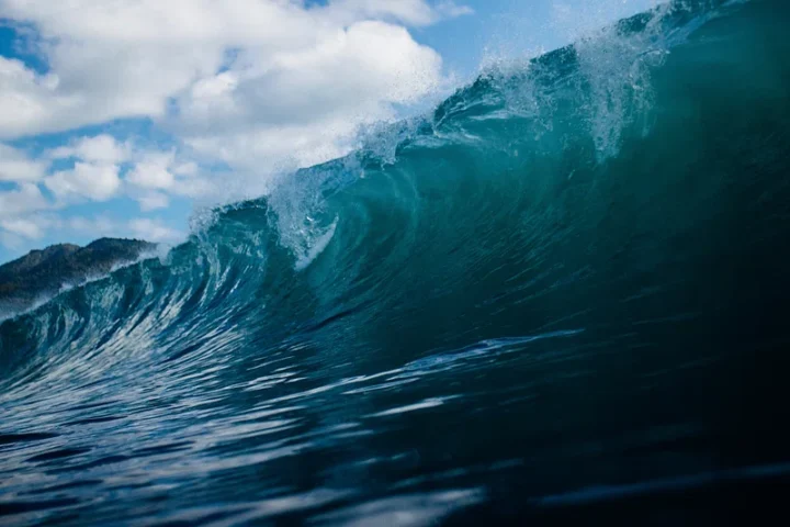Close-up perspective of a large turquoise ocean wave curling with white foam against a blue sky with clouds, demonstrating the immense power and scale of tsunami waves.