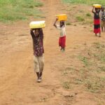Four children walk along a dusty path in rural Uganda, each balancing a yellow water container on their head.