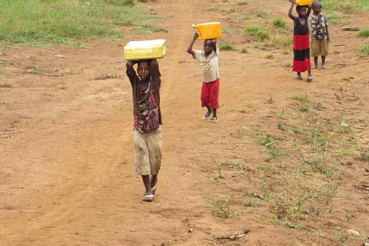 Four children walk along a dusty path in rural Uganda, each balancing a yellow water container on their head.