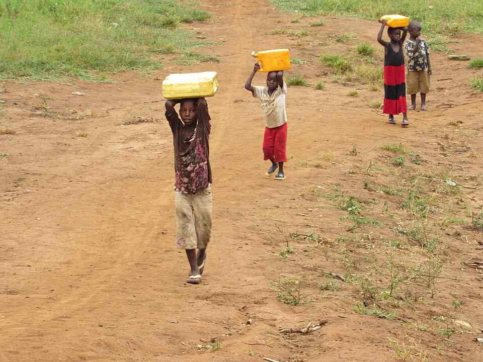 Four children walk along a dusty path in rural Uganda, each balancing a yellow water container on their head.