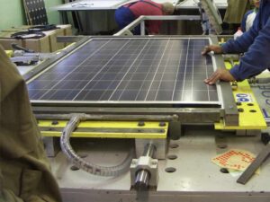 Workers operating a solar panel lamination machine in a manufacturing facility, placing a photovoltaic module into the laminator.