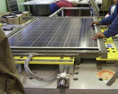 Workers operating a solar panel lamination machine in a manufacturing facility, placing a photovoltaic module into the laminator.
