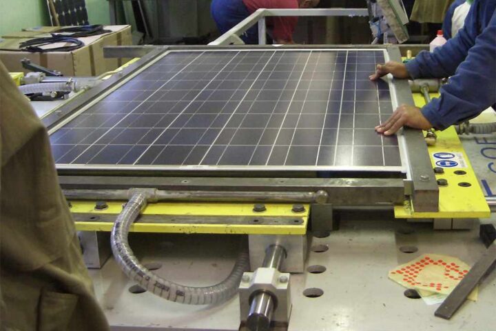 Workers operating a solar panel lamination machine in a manufacturing facility, placing a photovoltaic module into the laminator.