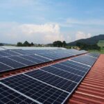 Solar panels installed on a red-tiled roof with mountains and countryside in the background under a blue sky with white clouds.