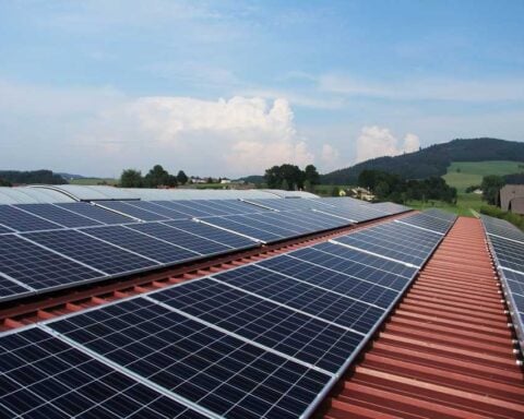 Solar panels installed on a red-tiled roof with mountains and countryside in the background under a blue sky with white clouds.