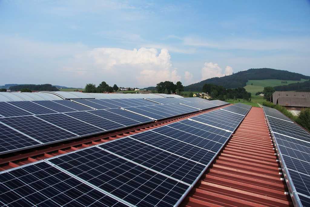 Solar panels installed on a red-tiled roof with mountains and countryside in the background under a blue sky with white clouds.