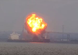 Flames and thick smoke billow from the bow of the W Sapphire bulk carrier near the Francis Scott Key Bridge as emergency vessels respond on Baltimore’s Patapsco River at dusk.