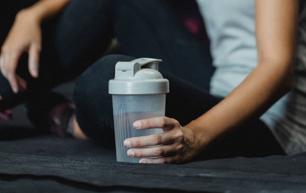 person in athletic wear holding a shaker bottle while seated on dark gym surface