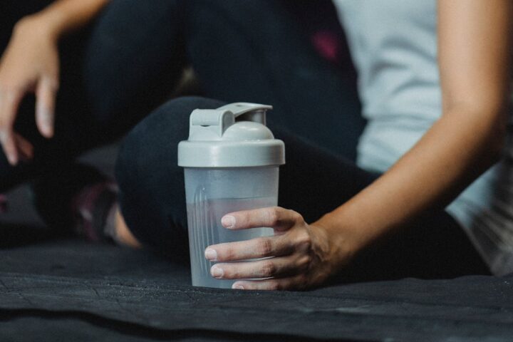 person in athletic wear holding a shaker bottle while seated on dark gym surface