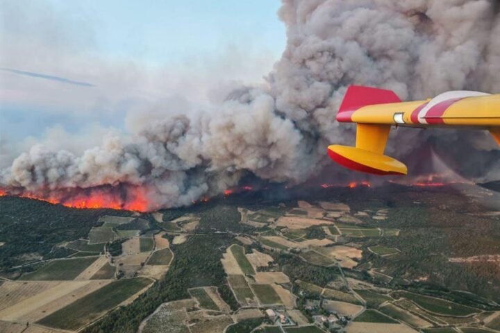 Canadair water bomber releasing a stream of retardant over rolling vineyards and scrubland as thick smoke plumes rise into a clear sky.
