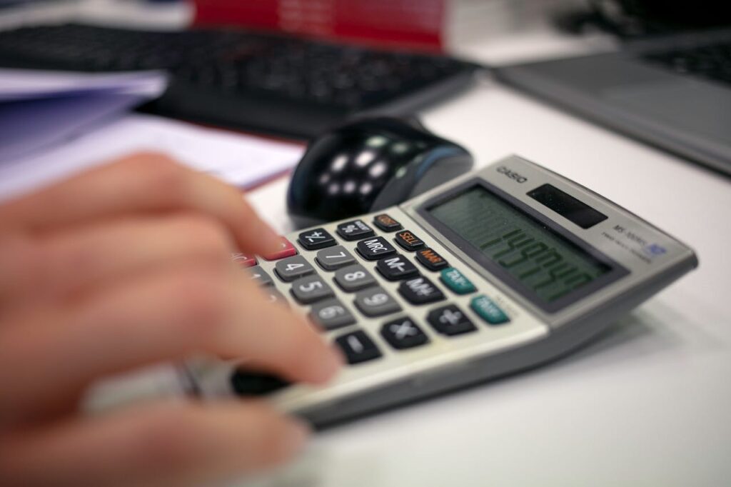 Close-up of a person's hand using a Casio calculator on a desk with computer peripherals visible in the background, demonstrating financial calculation in progress.