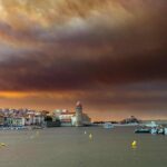 Wildfire smoke drifting over a Mediterranean coastal town with hills and sea under hazy skies, southern Europe, August 2025.