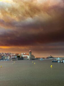 Wildfire smoke drifting over a Mediterranean coastal town with hills and sea under hazy skies, southern Europe, August 2025.