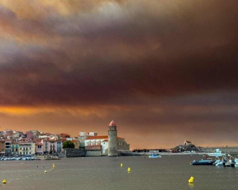 Wildfire smoke drifting over a Mediterranean coastal town with hills and sea under hazy skies, southern Europe, August 2025.