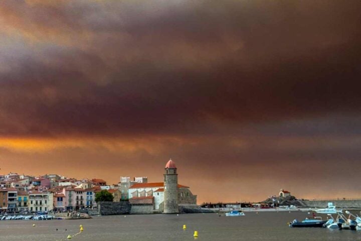 Wildfire smoke drifting over a Mediterranean coastal town with hills and sea under hazy skies, southern Europe, August 2025.