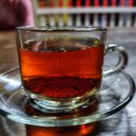 A clear glass tea cup filled with dark amber tea sits on a matching saucer, with blurred shelves in the background