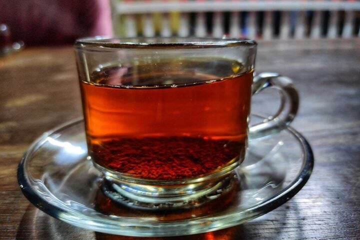 A clear glass tea cup filled with dark amber tea sits on a matching saucer, with blurred shelves in the background