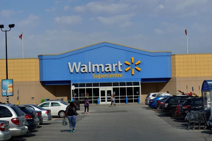 Exterior of a Walmart Supercentre store with blue and yellow signage, parked cars in the foreground, and shoppers walking toward the entrance.